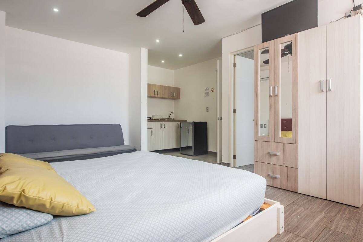 A bedroom with a bed, a wardrobe, and a kitchenette in the background.