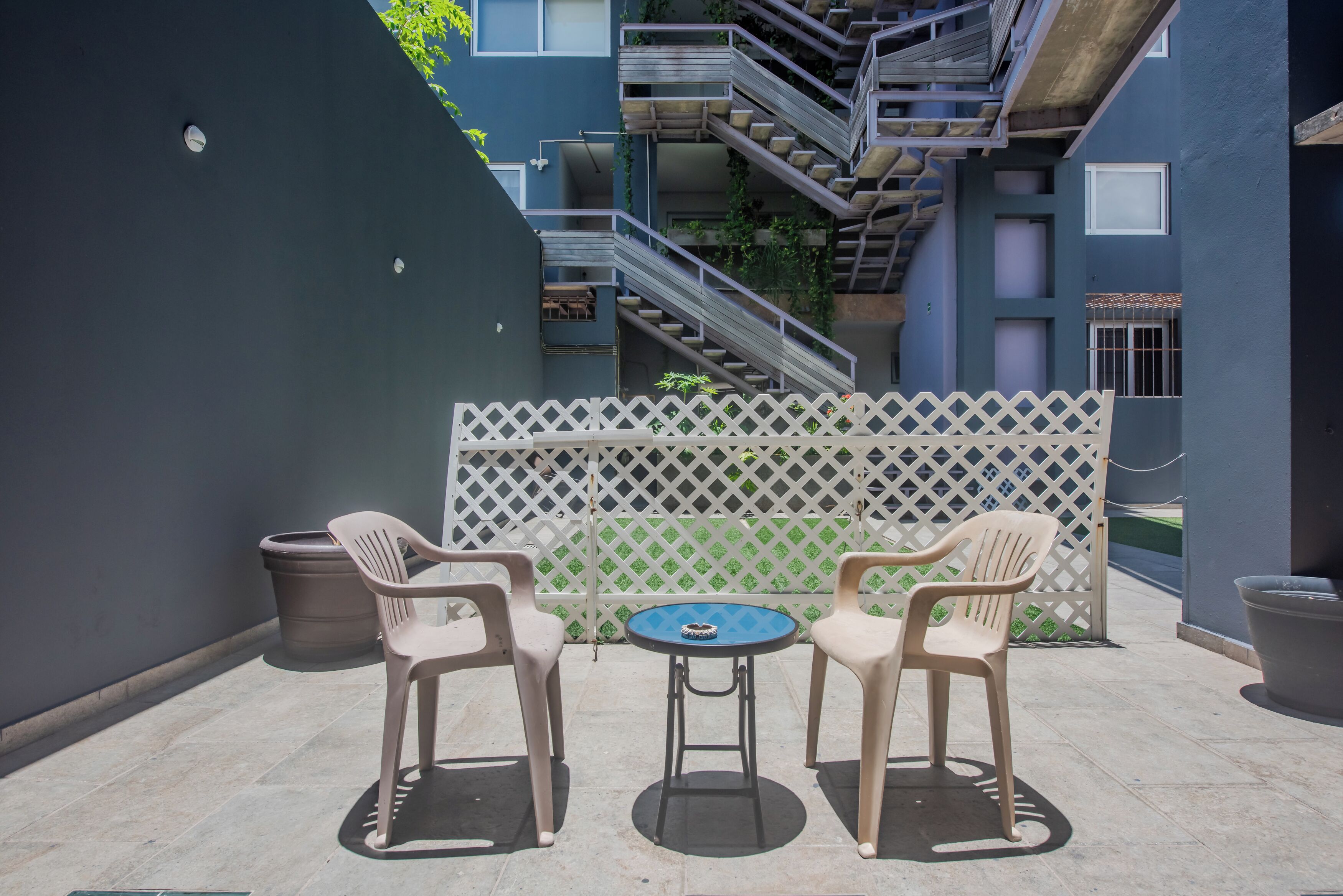 Un patio con una mesa y sillas, cercado por un enrejado blanco, y una escalera que conduce a un nivel superior.