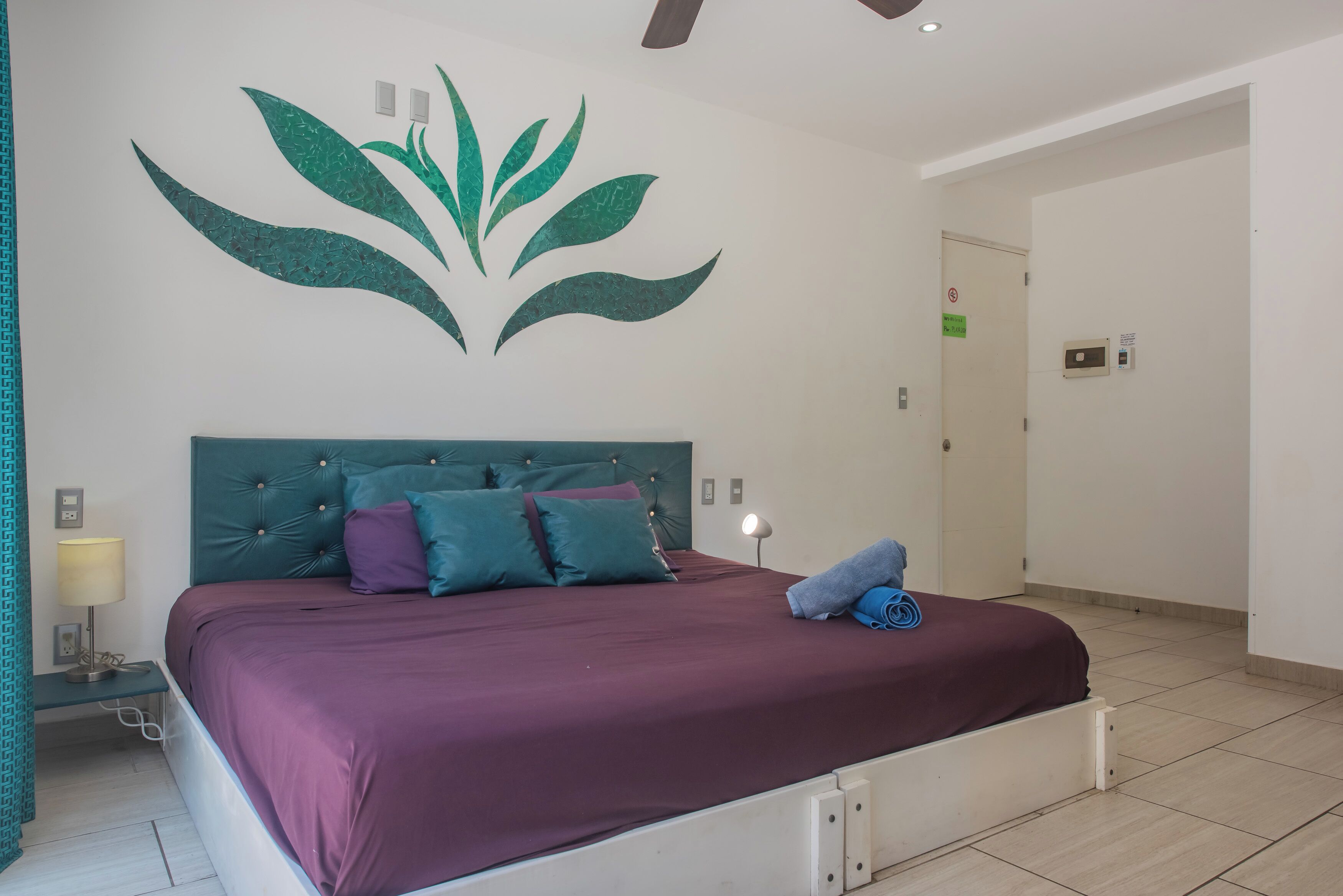 A bedroom with a bed, a wall-mounted light, and a green wall decoration.