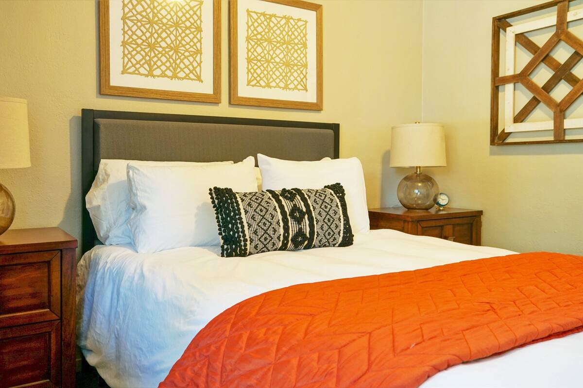 A neatly made bed with white linens and an orange blanket, flanked by two bedside tables with lamps, and two framed wall art pieces above.