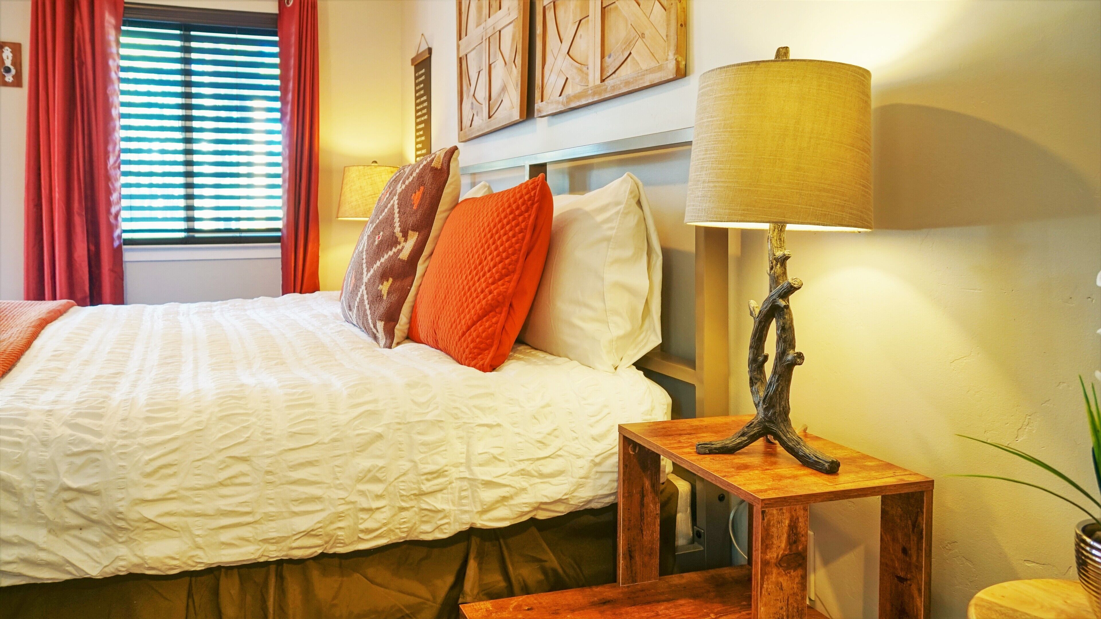 A neatly made bed with white and orange bedding, a wooden nightstand with a lamp, and a window with red curtains.