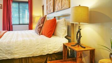 A neatly made bed with white and orange bedding, a wooden nightstand with a lamp, and a window with red curtains.