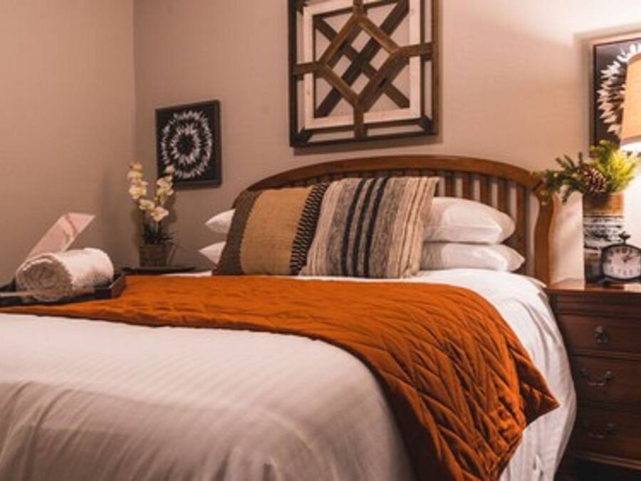 A neatly made bed with a white comforter and an orange throw blanket. There is a wooden headboard, a nightstand with a lamp, and a small clock.