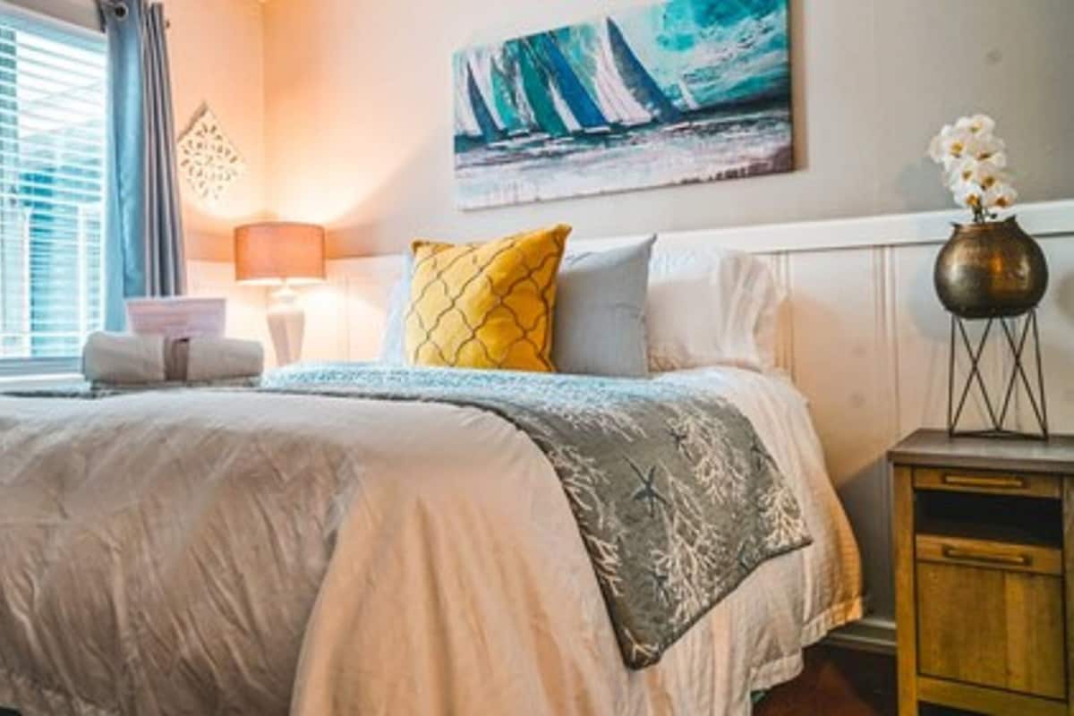 A neatly made bed with a white duvet, a yellow pillow, and a grey throw. There is a painting of sailboats on the wall, a lamp on a bedside table, and a vase with flowers on a nightstand.