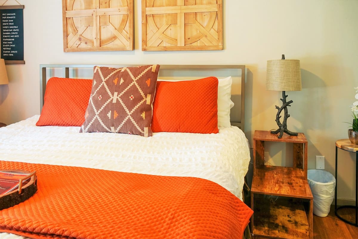 A neatly made bed with orange bedding, a patterned pillow, and a tray with a book. There are two wooden nightstands with lamps and a small potted plant.