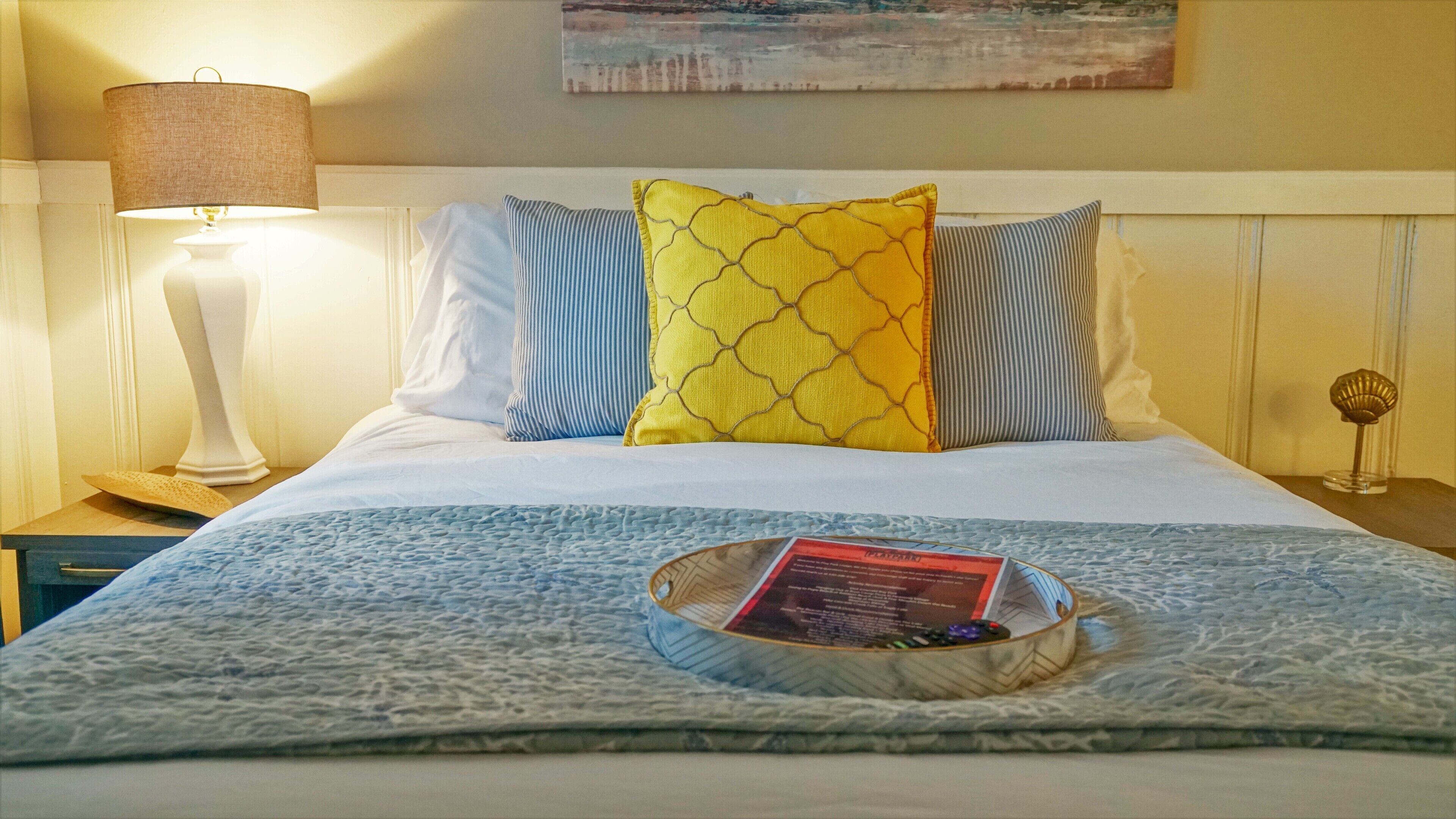 A neatly made bed with a patterned bedspread, two pillows, a bedside lamp, and a tray with a book and pen.