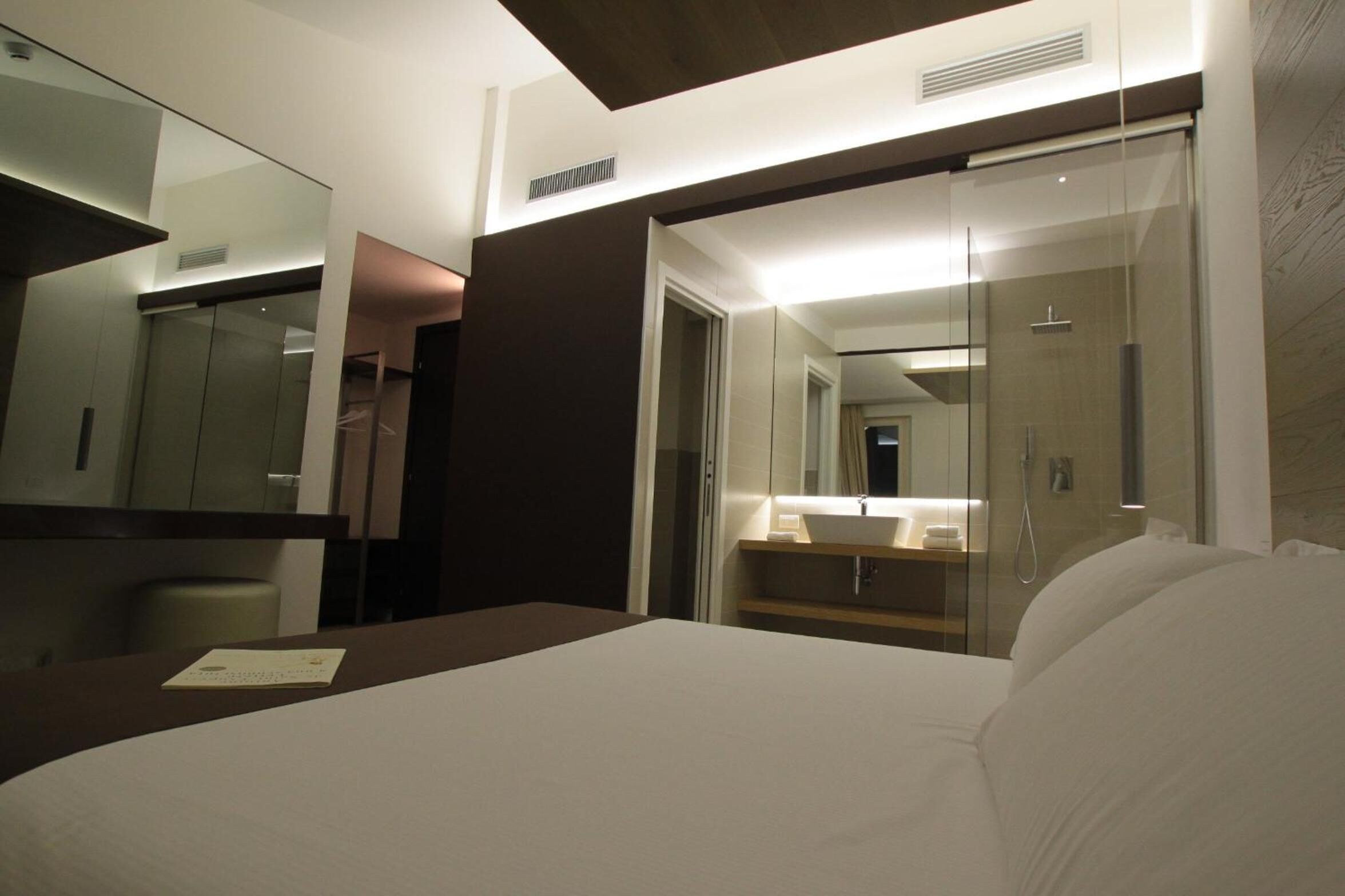 Habitación de hotel con cama, mesita de noche, espejo, armario y baño con lavabo y ducha.