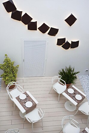A patio area with two tables set for dining, surrounded by white chairs. There are decorative wall lights and potted plants.