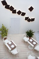 Un patio con dos mesas dispuestas para comer, rodeado de sillas blancas. Hay luces decorativas en la pared y plantas en macetas.