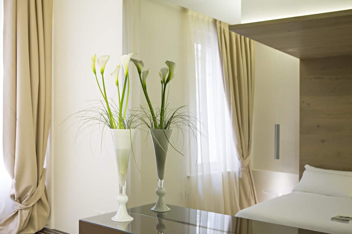 A hotel room with a bed, a bedside table with a vase of flowers, and a window with curtains.