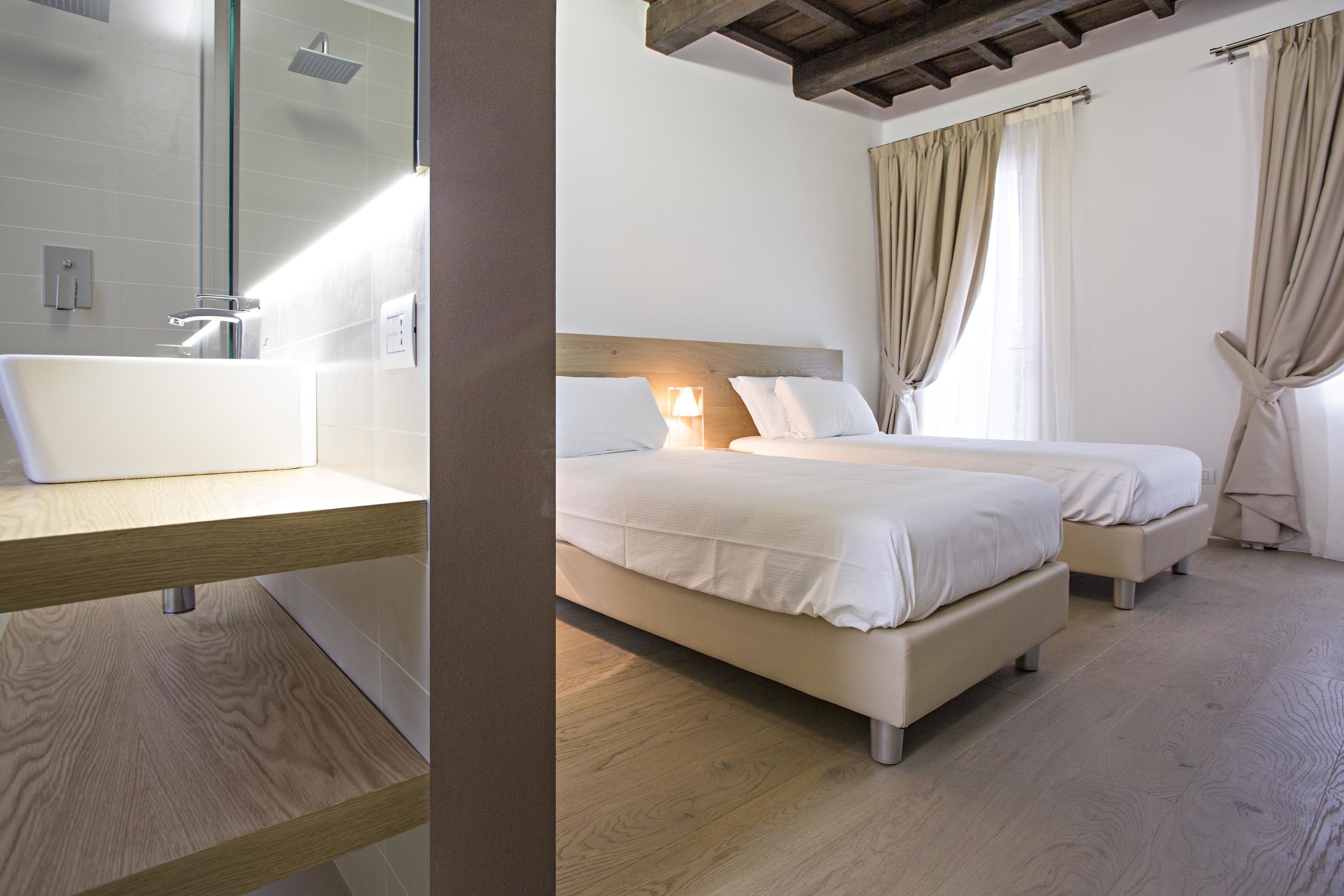 Ein modernes Hotelzimmer mit Holzboden, einem großen Spiegel, einem weißen Waschbecken, zwei Betten mit weißer Bettwäsche und einem Fenster mit Vorhängen.