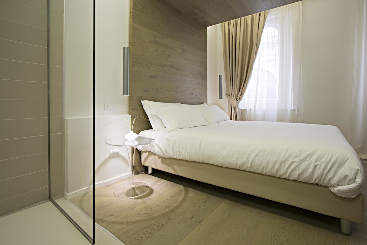 A modern bedroom with a wooden headboard, a bed with white bedding, a glass shower enclosure, and a bedside table with a vase.