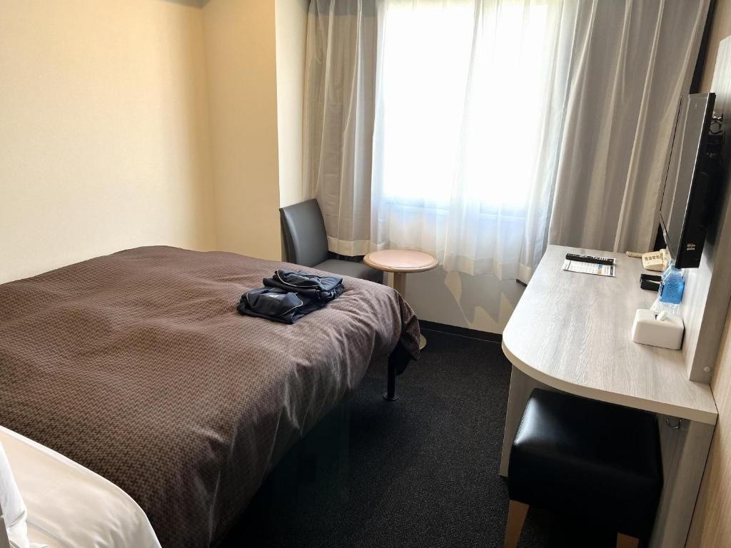 A hotel room with a bed, a desk, a chair, a small round table, a television, and a window with curtains.