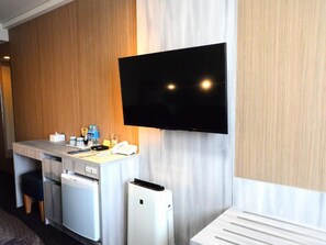 A hotel room with a flat-screen TV mounted on the wall, a small kitchenette with a refrigerator, and a white bedside table. - Sunwest Hotel Sasebo (Sasebo)