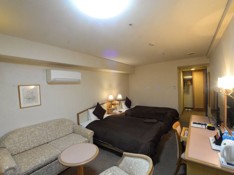 A hotel room with a sofa, two beds, a desk with a lamp, and a mirror.