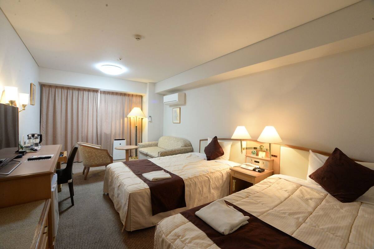 A hotel room with two beds, a desk, a chair, a lamp, and a window with curtains.