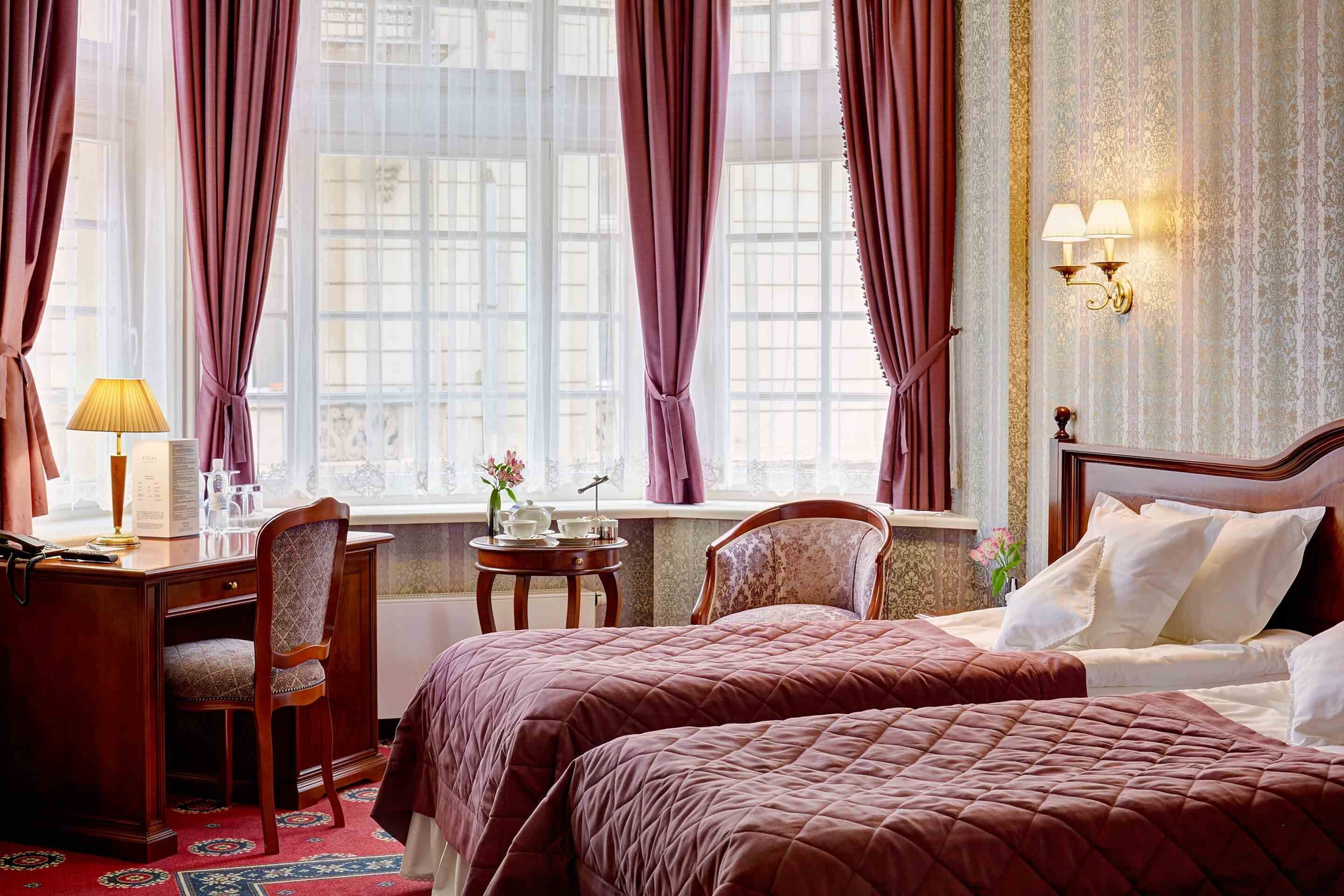A hotel room with two beds, a desk, a chair, a small table with a lamp, and a window with curtains.