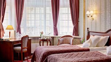 A hotel room with two beds, a desk, a chair, a small table with a lamp, and a window with curtains.