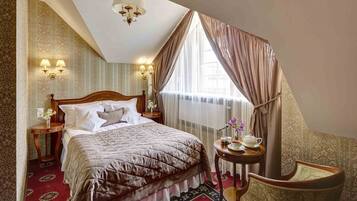 Habitación de hotel con una cama grande, una mesita de noche con lámpara, una silla y un ventanal con cortinas.