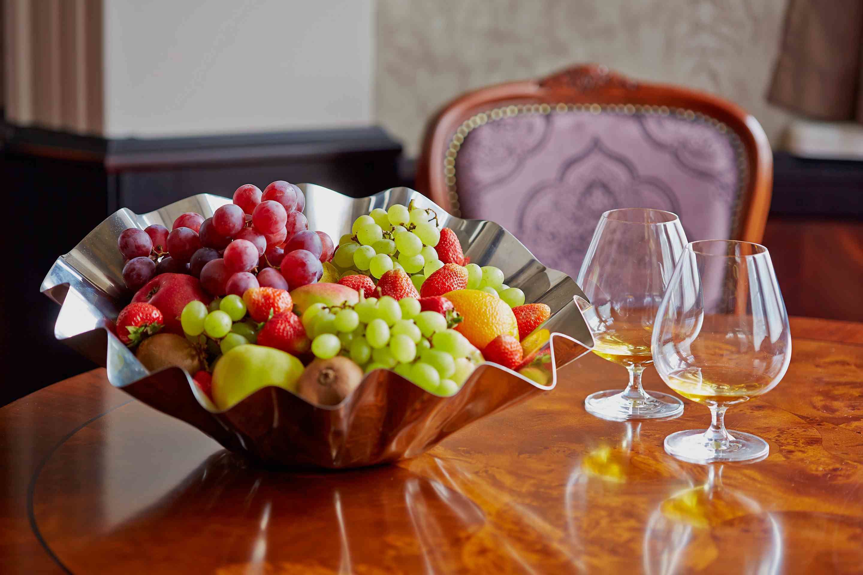 Un frutero con uvas, fresas y otras frutas sobre una mesa de madera, acompañado por dos copas de vino.