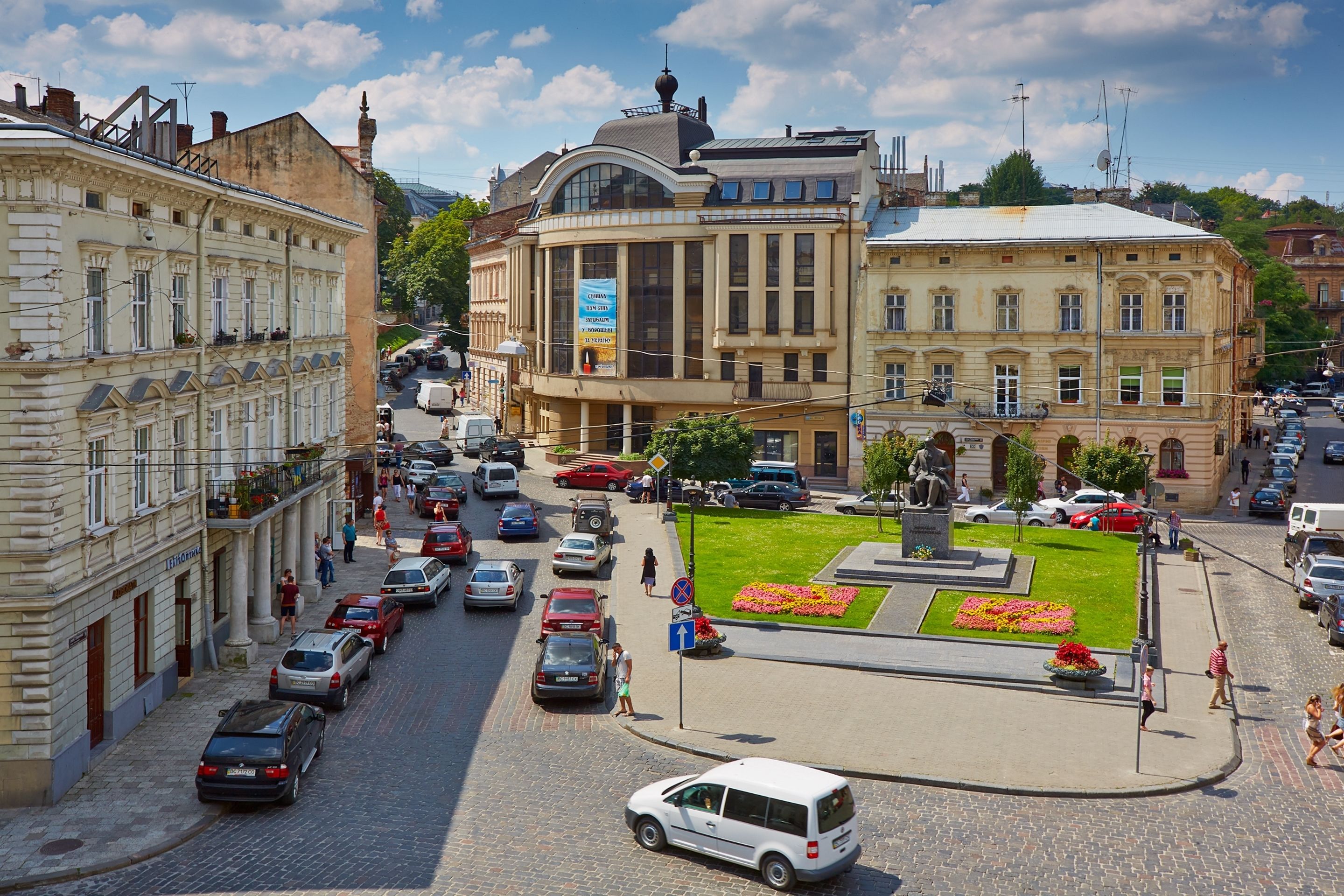 Un edificio histórico con arquitectura clásica, una plaza central con una estatua y una calle adoquinada con automóviles estacionados.