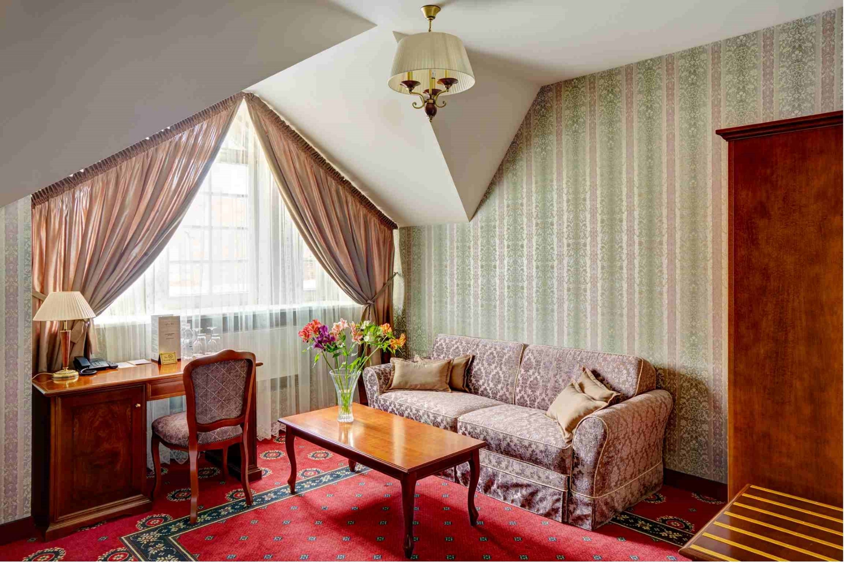 A room with a sofa, a desk with a lamp, a chair, a coffee table, and a vase with flowers.
