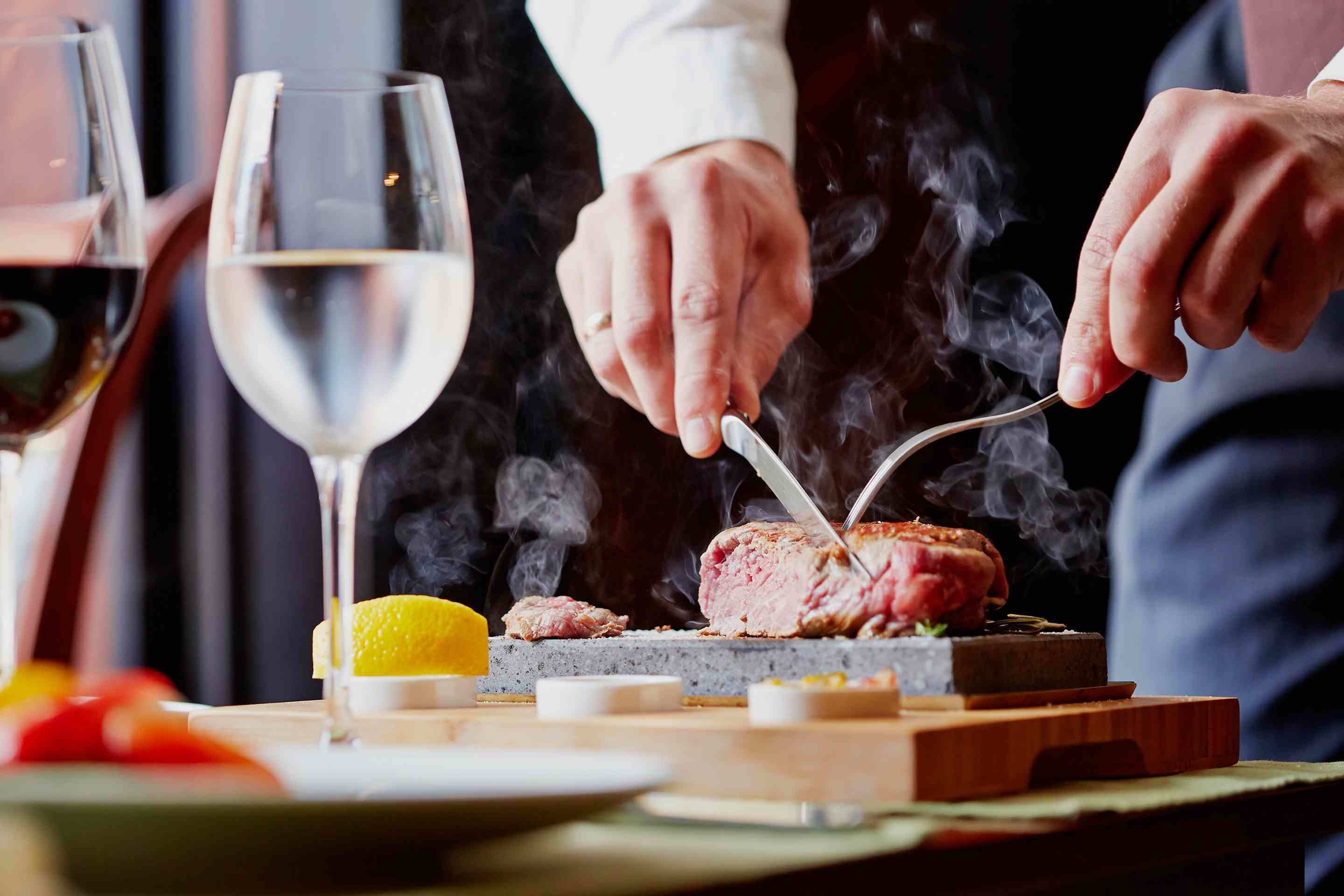 A person carving a steak on a grill with a knife and fork, with a glass of wine and a lemon on the table.