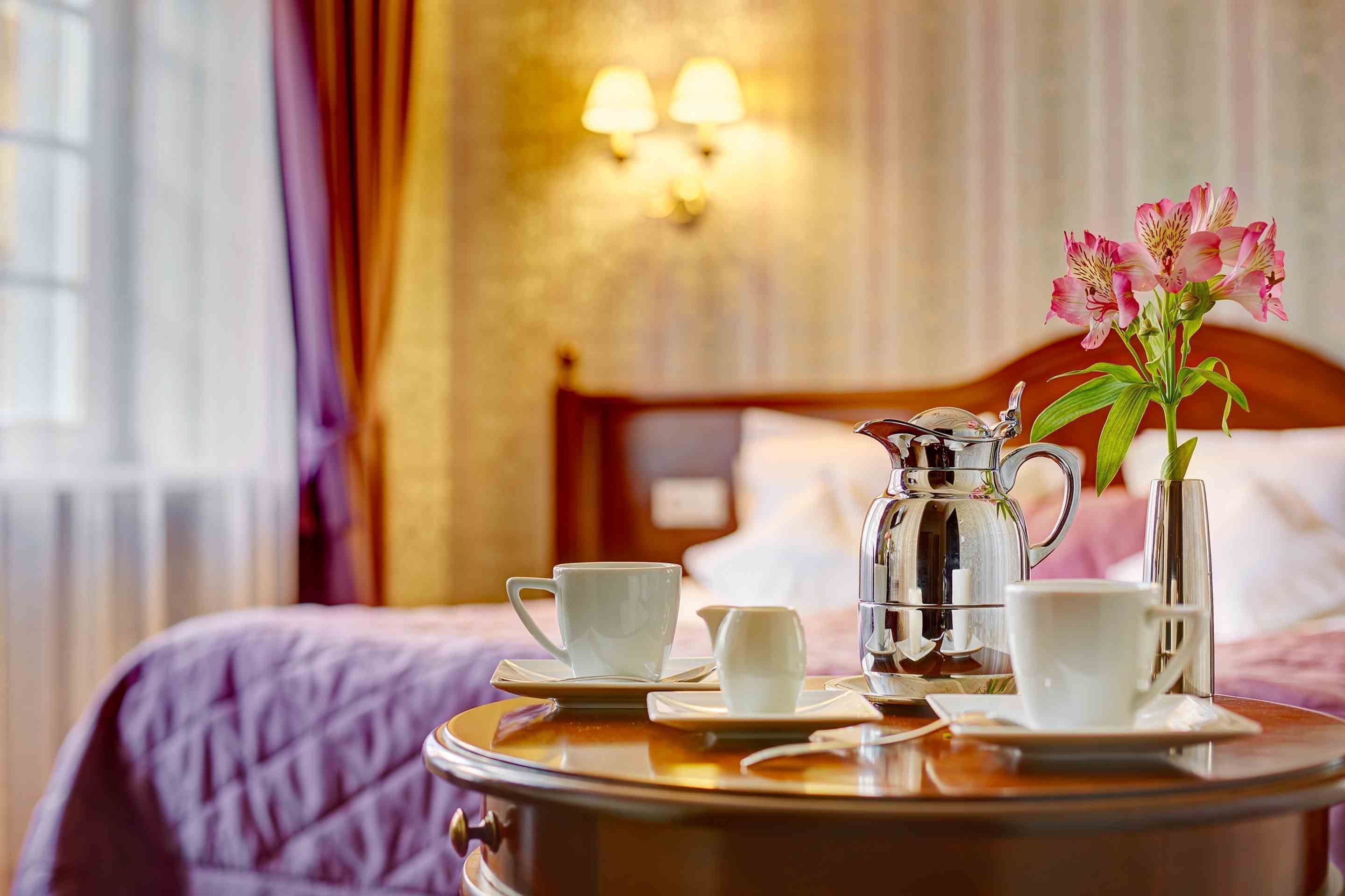A hotel room with a bed, a bedside table with a coffee pot, cups, and a vase with flowers.