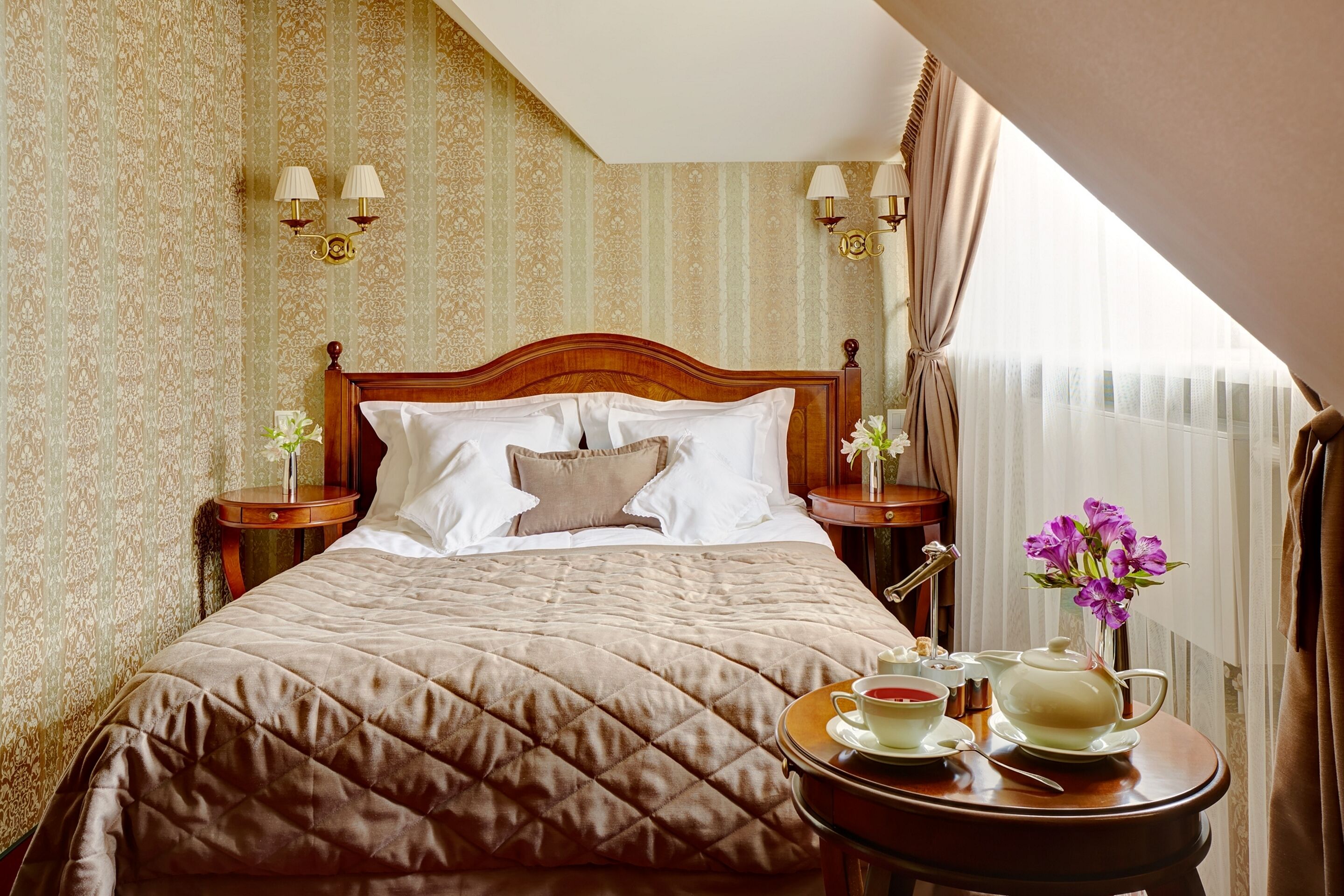 A neatly made bed with a quilted cover, two bedside tables with lamps, a tray with tea and a teapot, and a window with curtains.