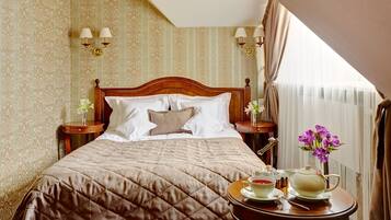 A neatly made bed with a quilted cover, two bedside tables with lamps, a tray with tea and a teapot, and a window with curtains.