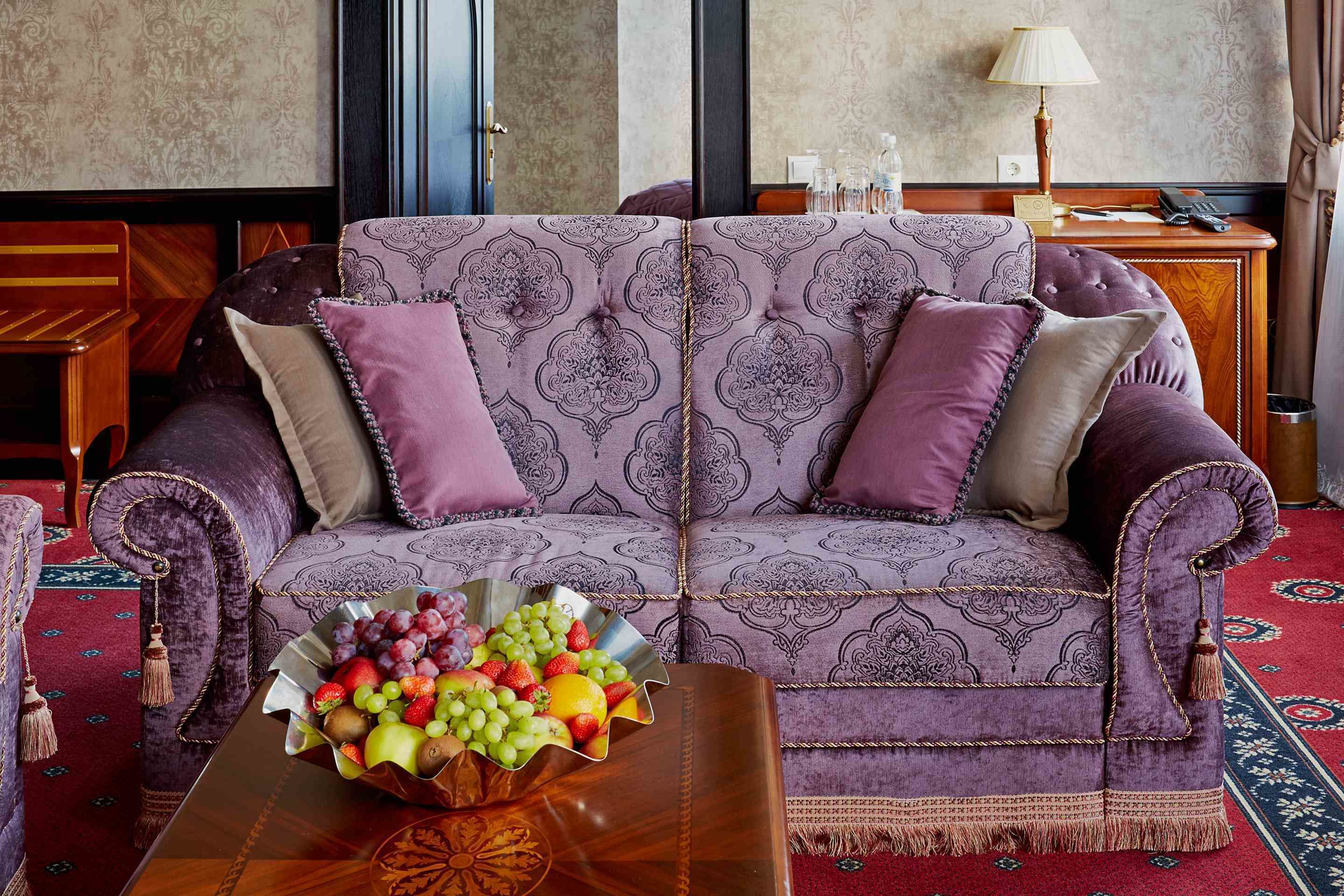Un sofá morado y mullido con cojines estampados, un frutero sobre una mesita de madera y una alfombra roja con diseño.