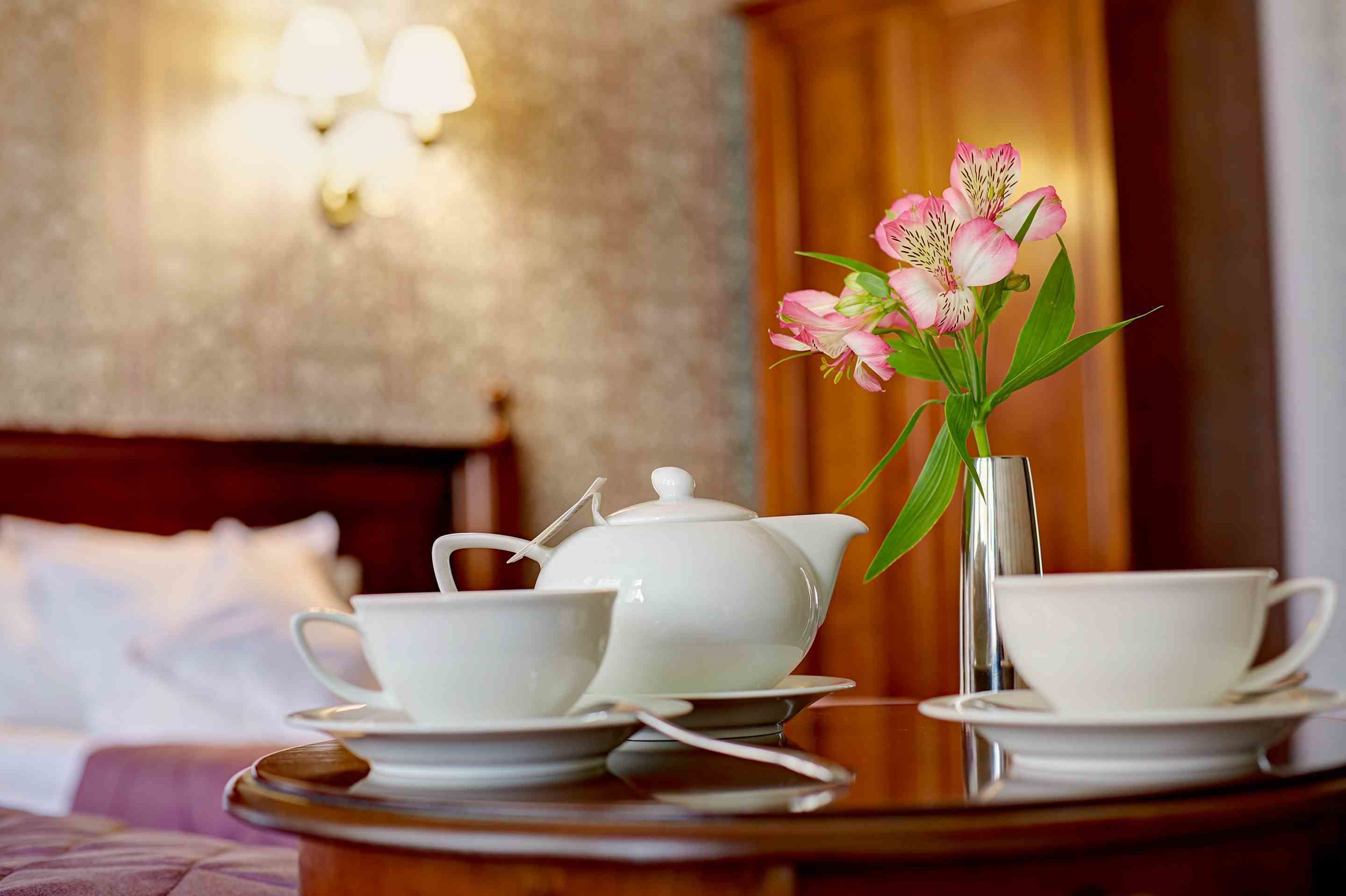 A neatly arranged tea set on a wooden table with a vase of pink flowers, in a room with a wooden headboard and wall sconces.