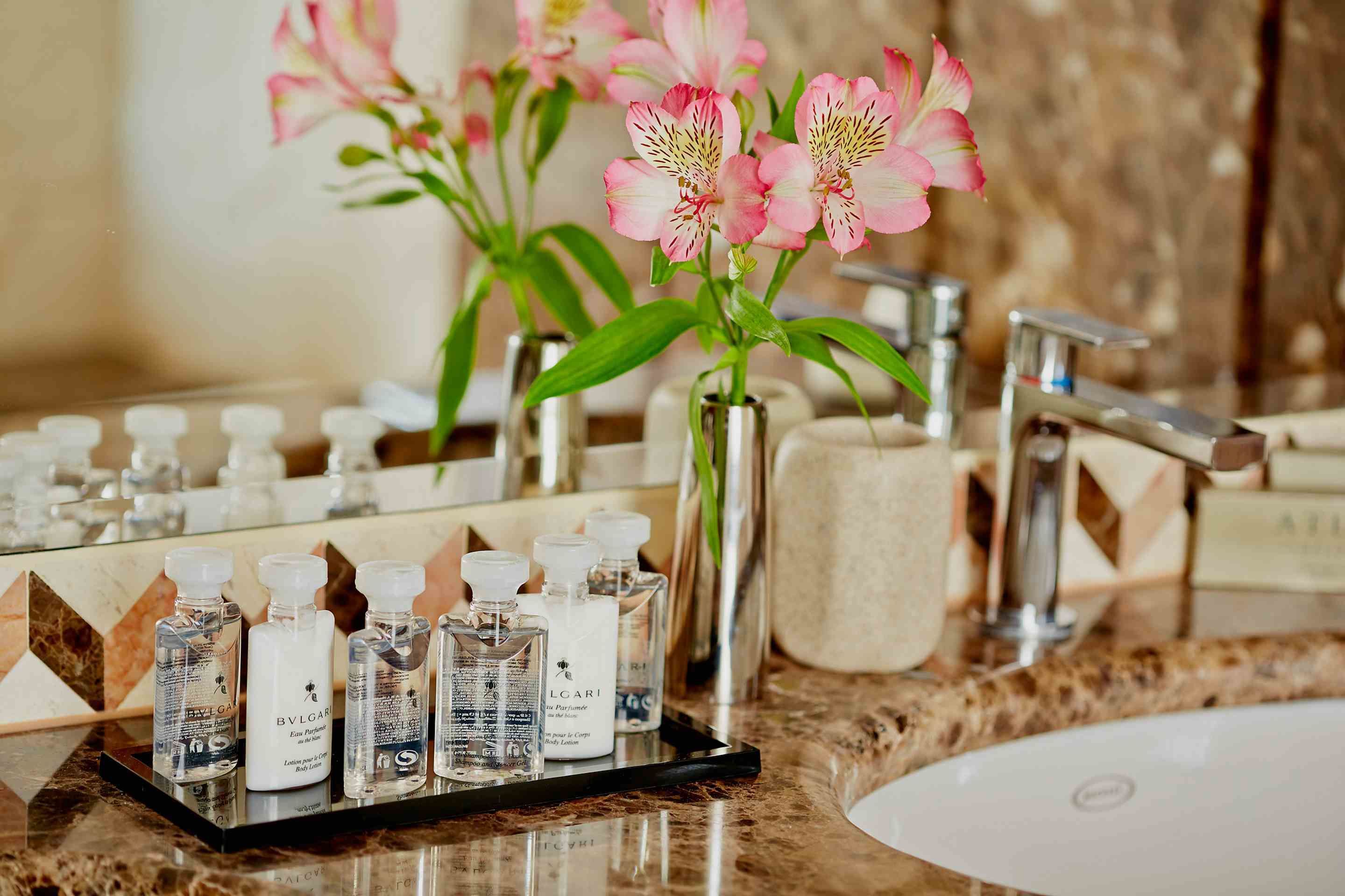A bathroom vanity with a marble countertop, a sink, and a mirror. There are several bottles of toiletries on the counter, including a white bottle labeled "BVLGARI" and a black bottle with a white cap.
