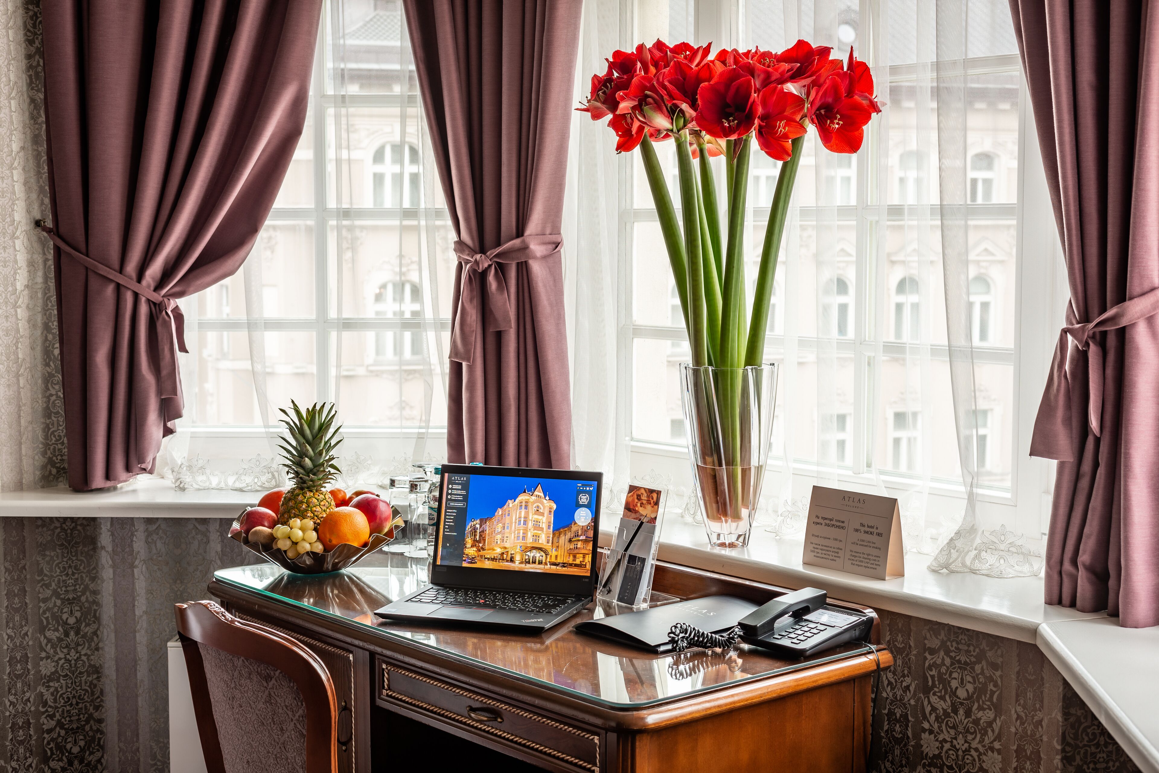 A desk with a laptop displaying a cityscape, a vase with red flowers, a fruit bowl, and a telephone.