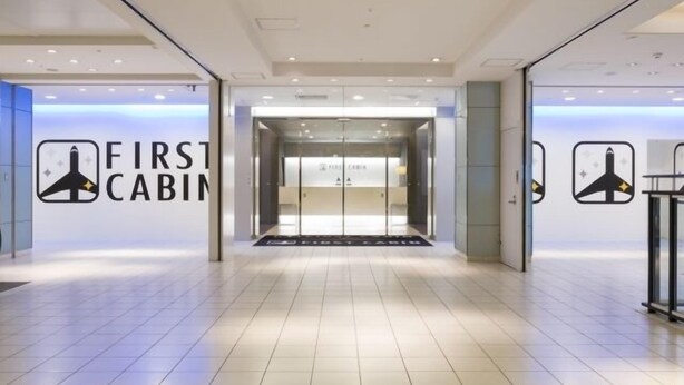 A modern hotel entrance with a glass door, a sign reading "FIRST CABIN," and a logo featuring a stylized airplane. - First Cabin Hakata (Fukuoka)