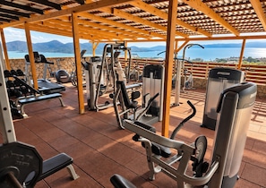 Un gimnasio al aire libre con diversos equipos de ejercicio, un área techada y una vista panorámica de un lago y montañas.