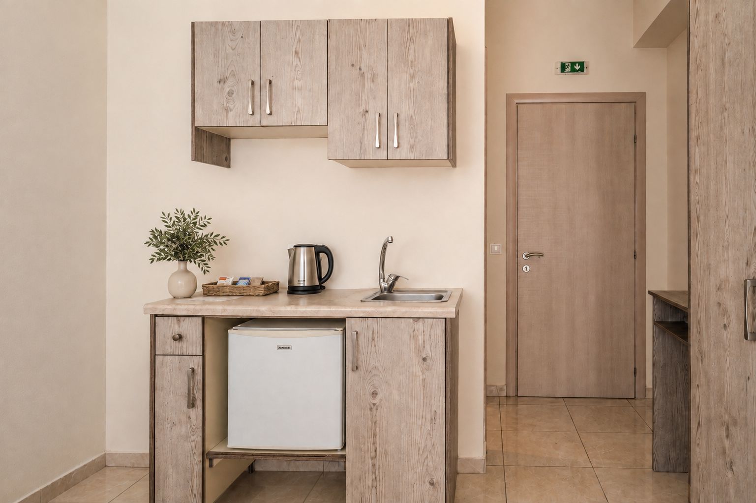 Une petite cuisine équipée d’un réfrigérateur encastré, d’un évier et d’une bouilloire. On peut voir une petite plante en pot sur le plan de travail.