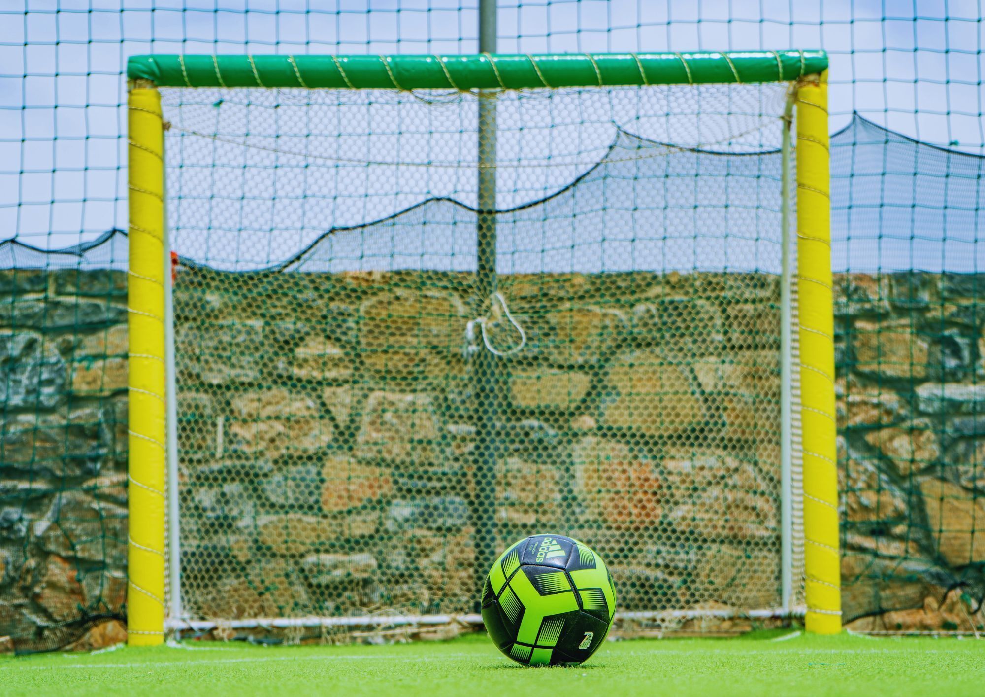 Una portería de fútbol con una red, una pelota y un muro de piedra al fondo.