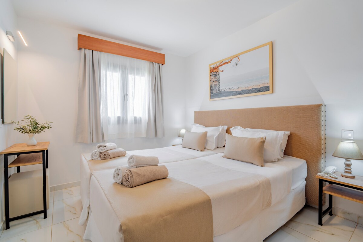 Ein Hotelzimmer mit zwei Betten, einem Bild an der Wand, einem Nachttisch mit Lampe und einem Fenster mit Vorhängen.