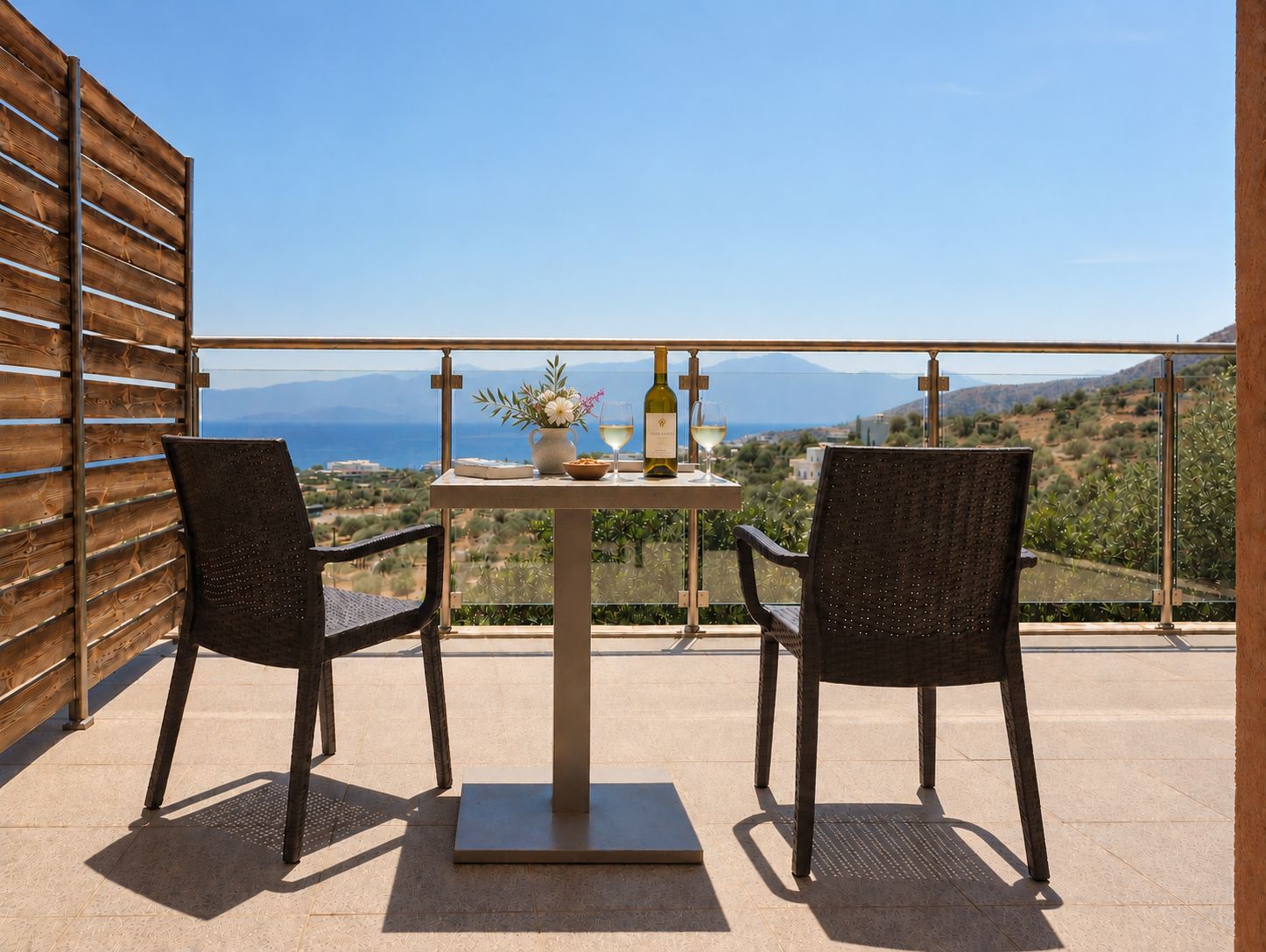 Varanda com duas cadeiras, uma mesa, uma garrafa e um copo, e vista para o mar e montanhas.