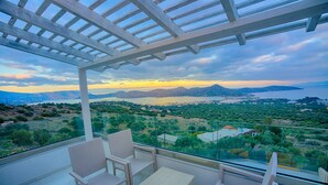 A balcony with wicker furniture and a glass railing overlooking a scenic landscape with mountains and a body of water.