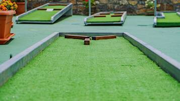 Una mesa de billar en miniatura con un obstáculo central, rodeada por un muro de piedra y plantas en macetas.
