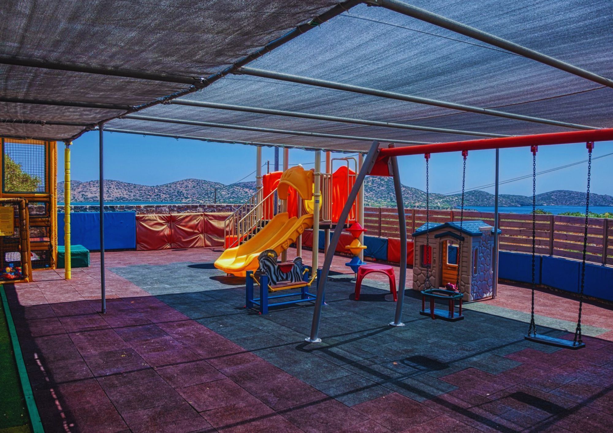 Ein Spielplatz mit Rutsche und Schaukeln, vor einer Bergkulisse und wolkenlosem Himmel.