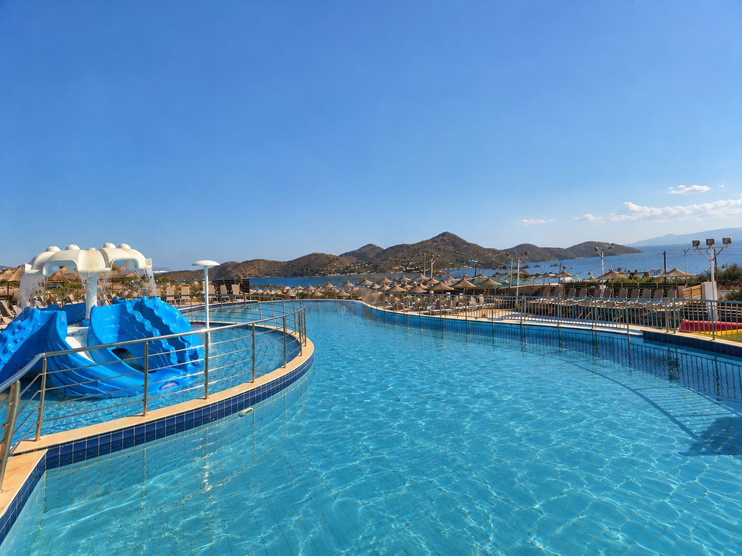 A swimming pool with a water slide, lounge chairs, umbrellas, and a mountainous backdrop.