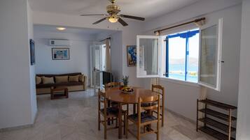 A dining area with a wooden table and chairs, a ceiling fan, and a window with a view of the sea.