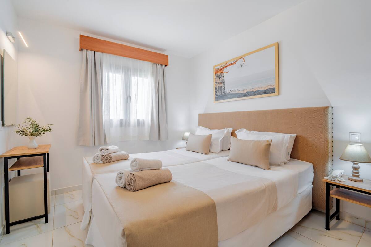 Ein Hotelzimmer mit zwei Betten, einem Bild an der Wand, einem Nachttisch mit Lampe und einem Fenster mit Vorhängen.