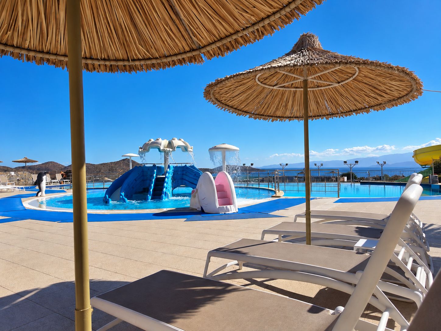 A pool area with lounge chairs, a water slide, and a clear view of the sea.