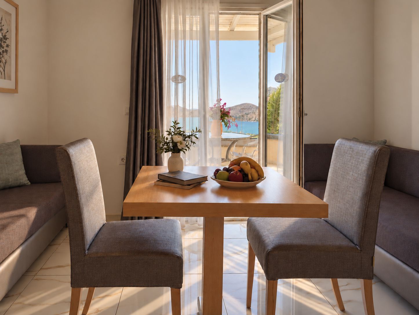 A room with a wooden table, two grey armchairs, a sofa, a vase with flowers, and a bowl of fruit.
