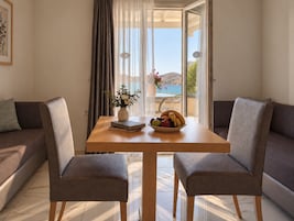 Una habitación con una mesa de madera, dos sillones grises, un sofá, un florero con flores y un cuenco de fruta.