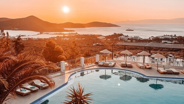 A resort pool area with lounge chairs and umbrellas, overlooking a coastal landscape with mountains and a body of water.
